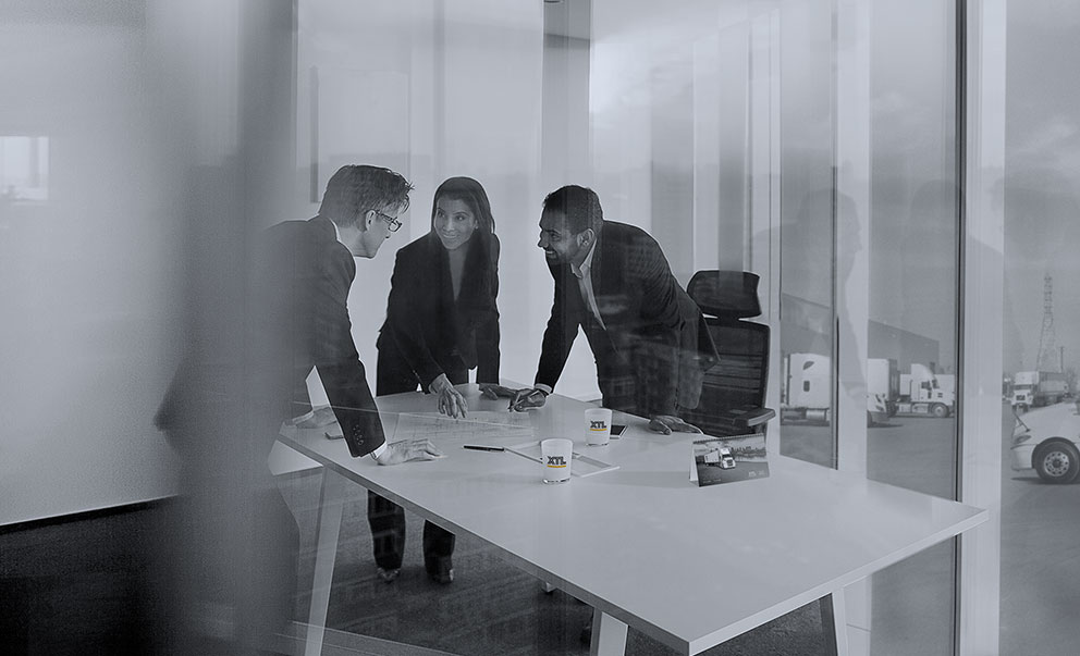 Three business people standing around XTL boardroom table with XTL trucks outside of the windows